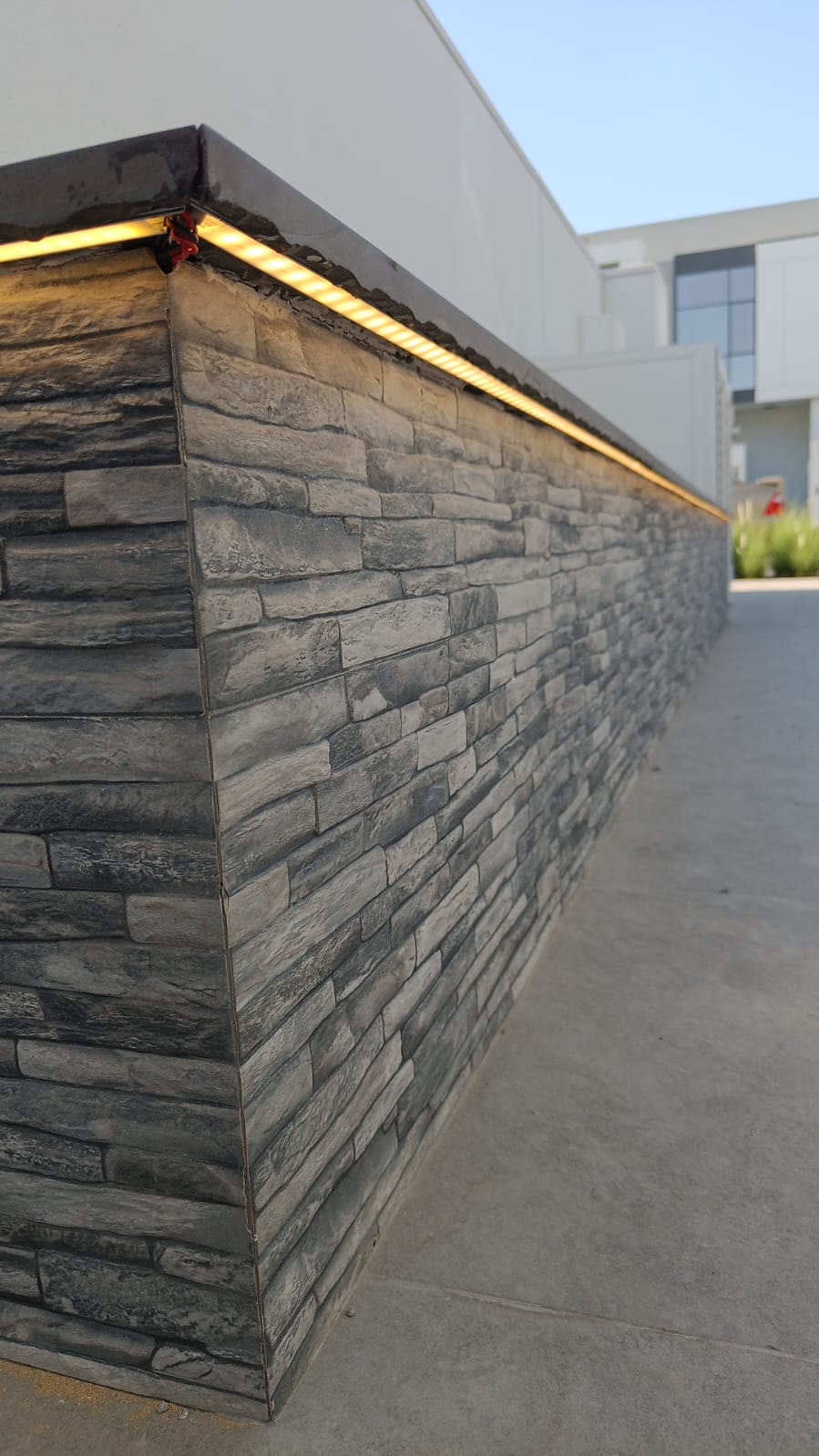 Low stone-clad wall with warm LED strip lighting under the dark capstone, beside a grey patio
